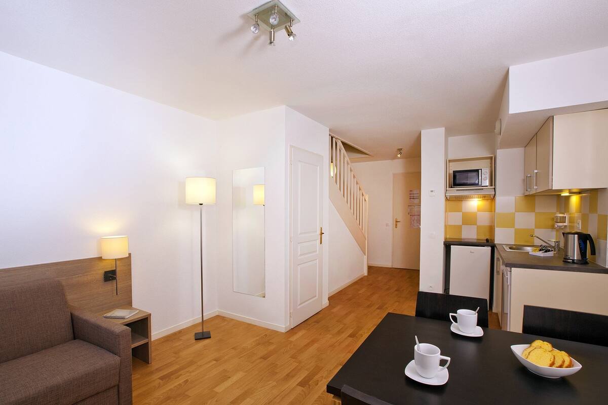 A compact living space with a sofa, bedside table, lamp, dining table with cups and a bowl, kitchenette, and a staircase.