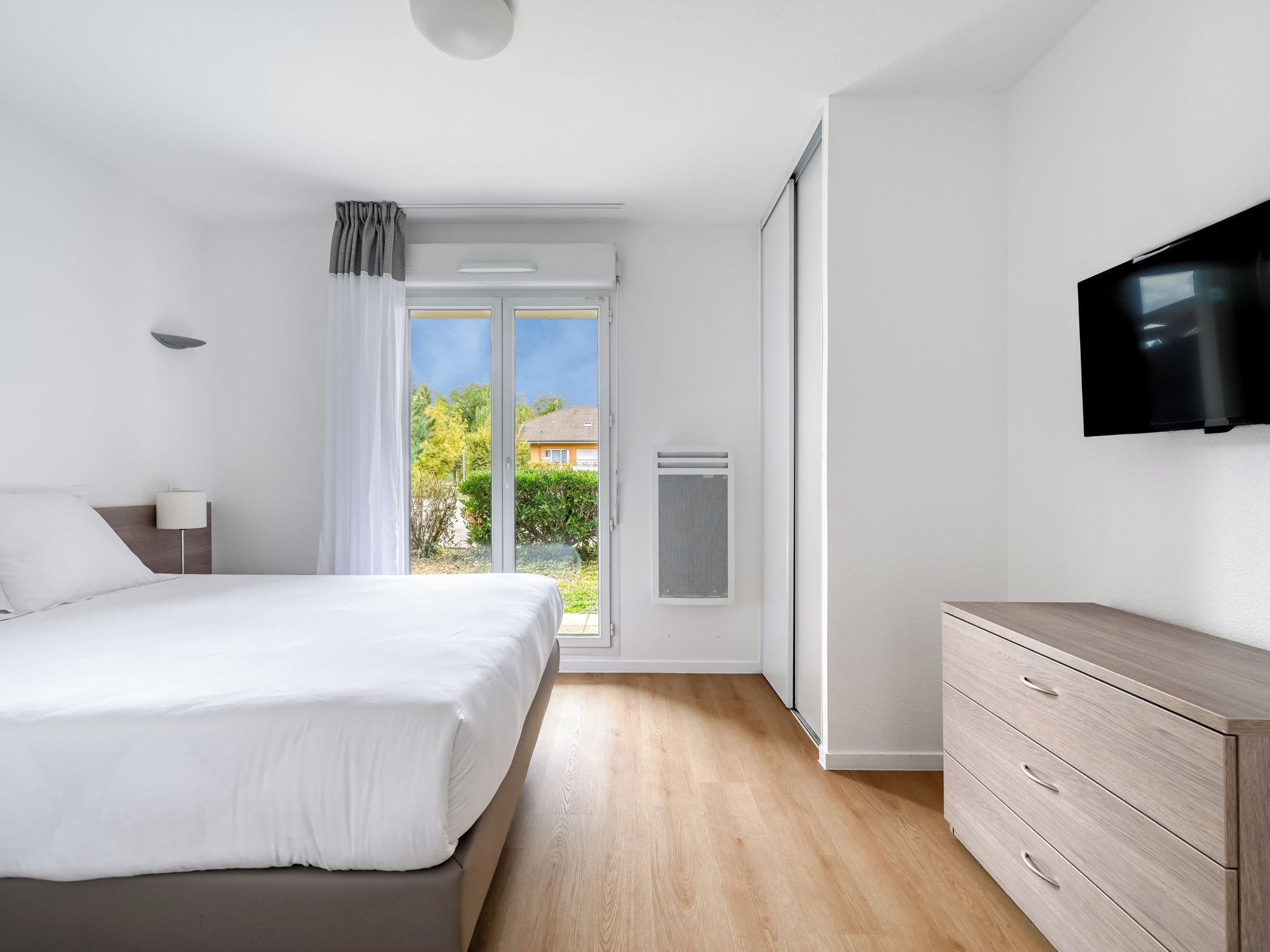 A modern bedroom with a bed, a dresser, a television, and a sliding glass door leading to a balcony.