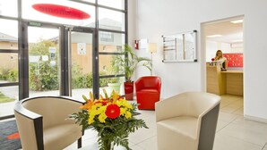 Un área de recepción con una silla roja, una silla color beige y un jarrón con flores.