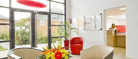Un área de recepción con una silla roja, una silla color beige y un jarrón con flores.