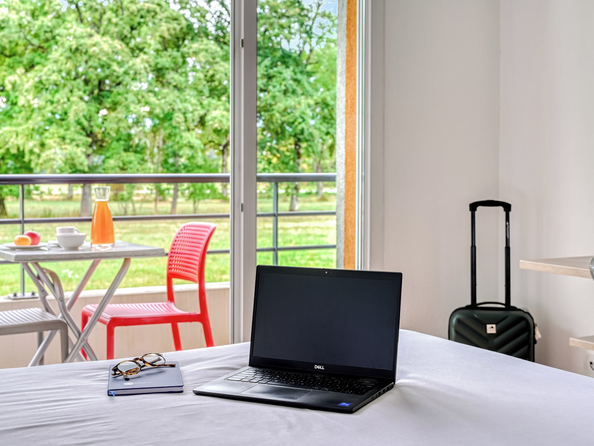 En bærbar computer på en seng med udsigt til en balkon, en kuffert og et bord med en drink.