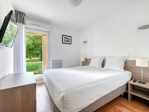 Quarto de hotel com cama, mesa de cabeceira, luminária e televisão sobre um suporte.