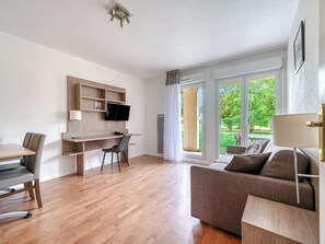Un salon moderne avec un plancher en bois, un canapé, un bureau avec une chaise, un téléviseur fixé au mur et une fenêtre avec des rideaux.