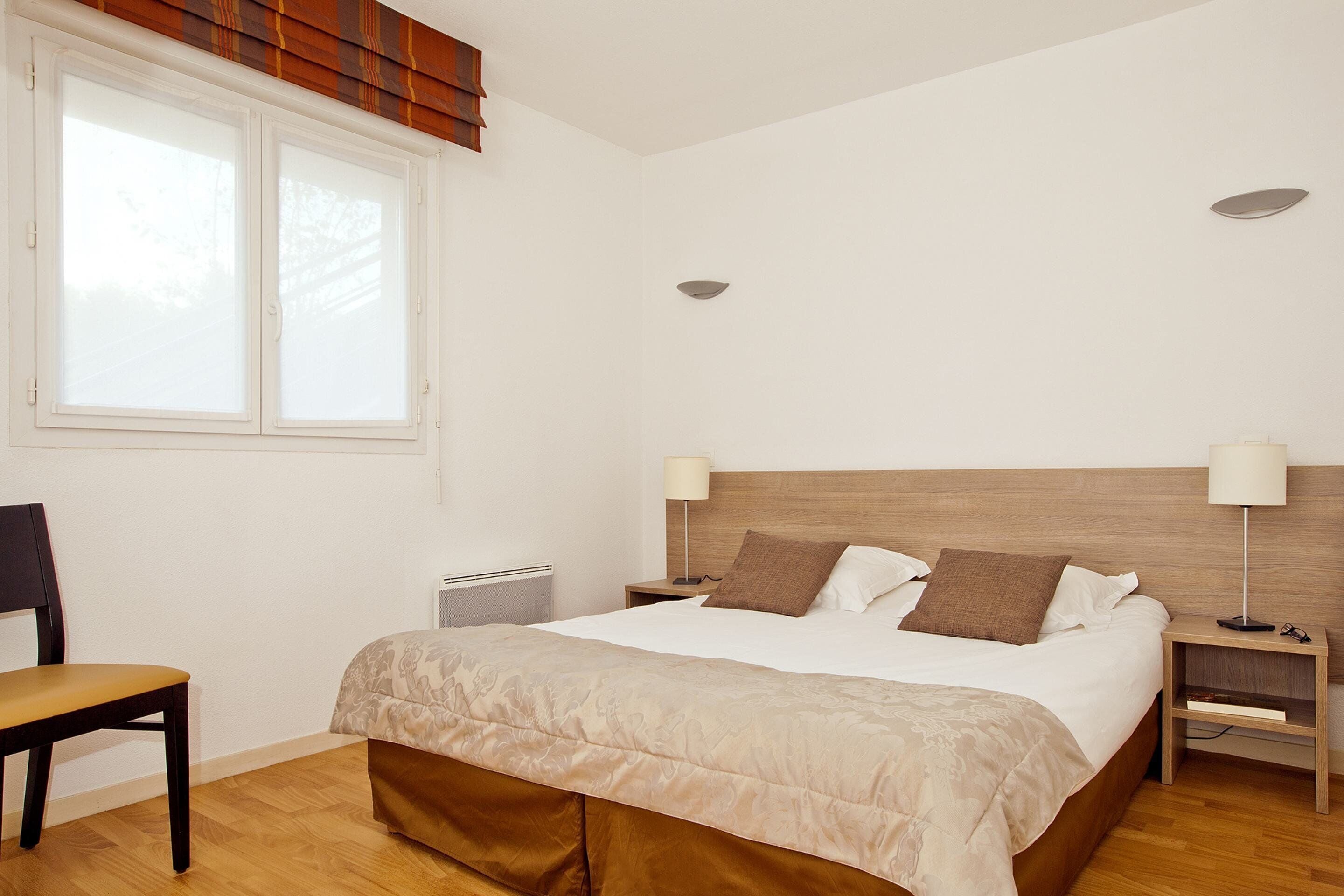 Une chambre à coucher comprenant un lit, deux tables de chevet, une chaise, une fenêtre et un radiateur.