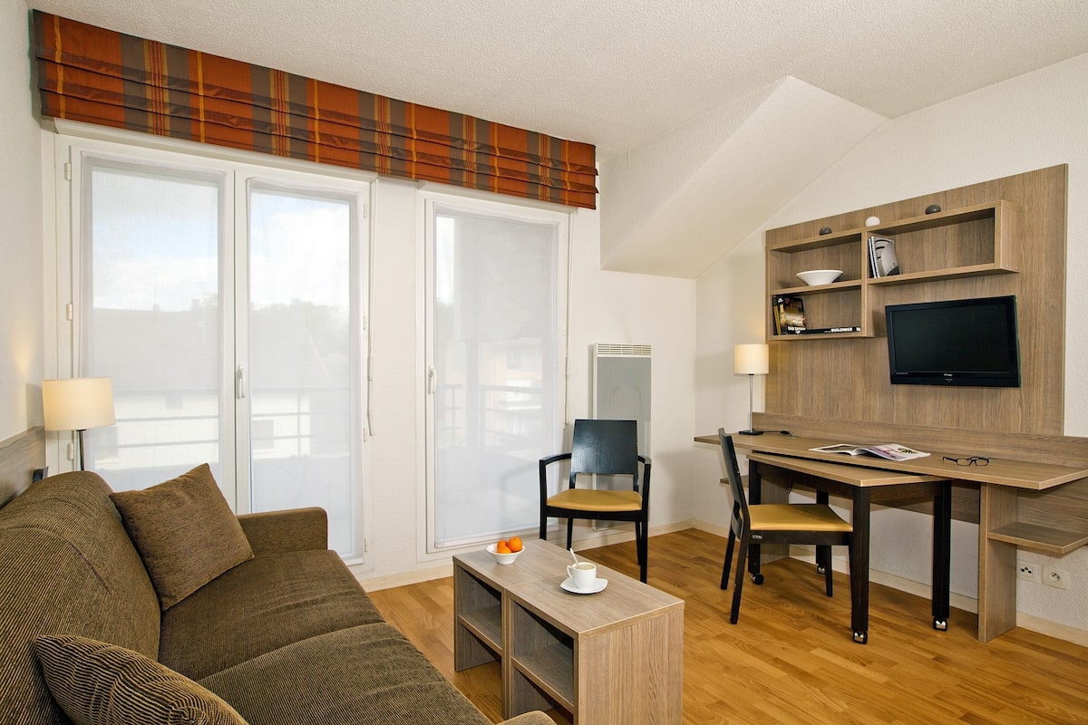 A modern living room with a sofa, a coffee table, a dining table with chairs, a television mounted on the wall, and a large window with a view.