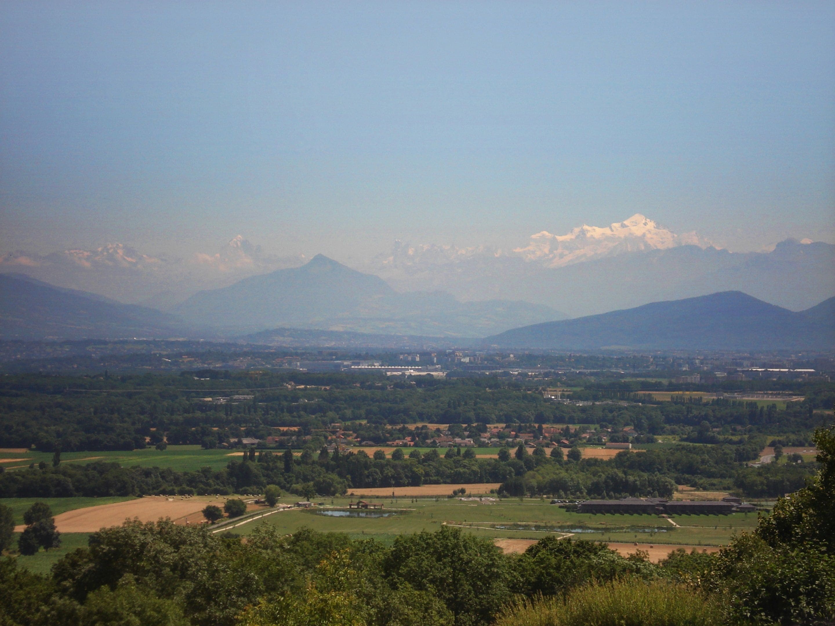 Una vista panorámica de un valle con montañas al fondo, campos verdes y edificios dispersos.