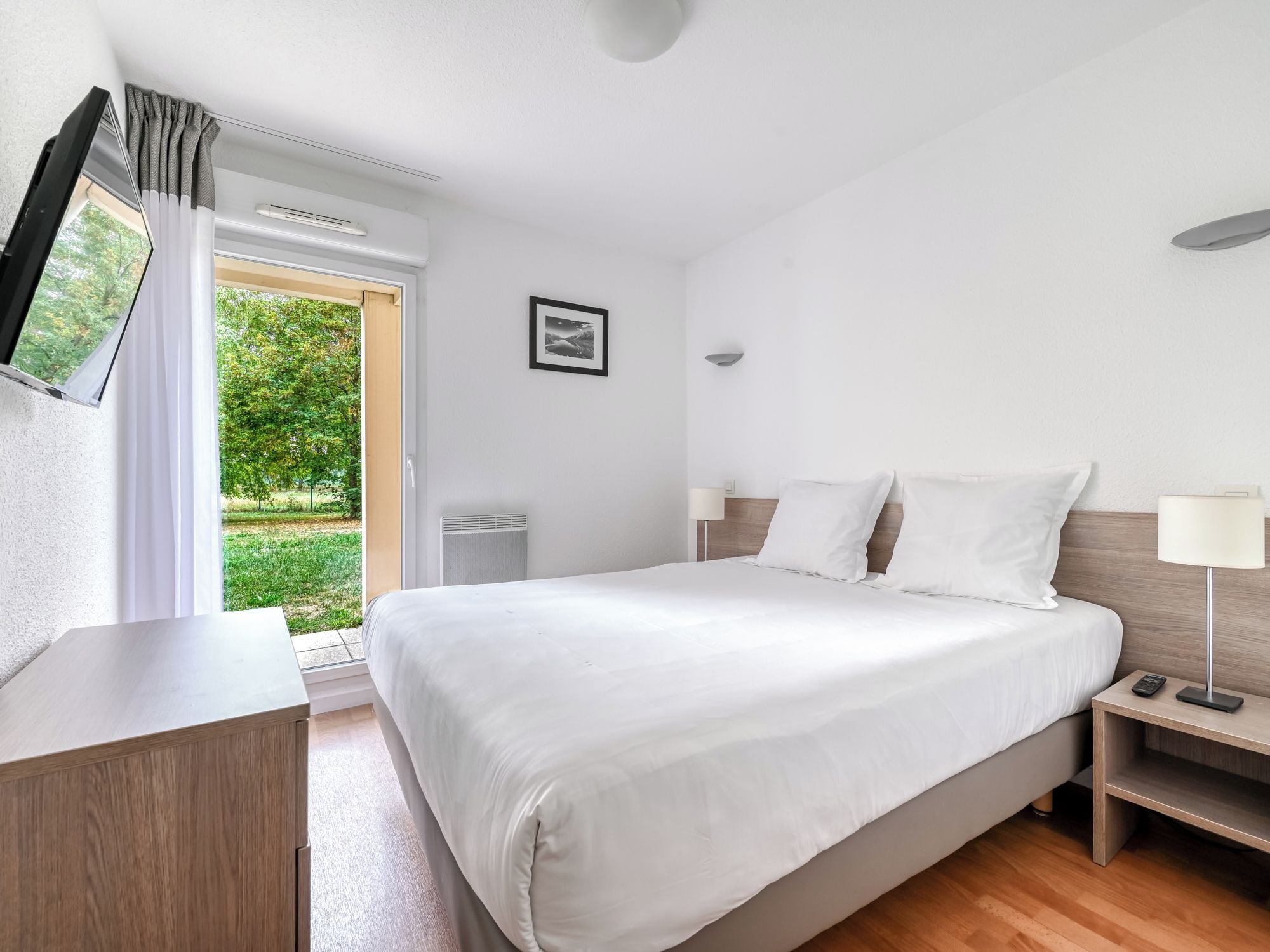 Una habitación de hotel con cama, mesita de noche, lámpara y televisor en la pared.