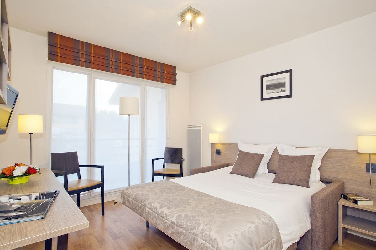 A hotel room with a large bed, two chairs, a desk with a lamp, and a window with a view.