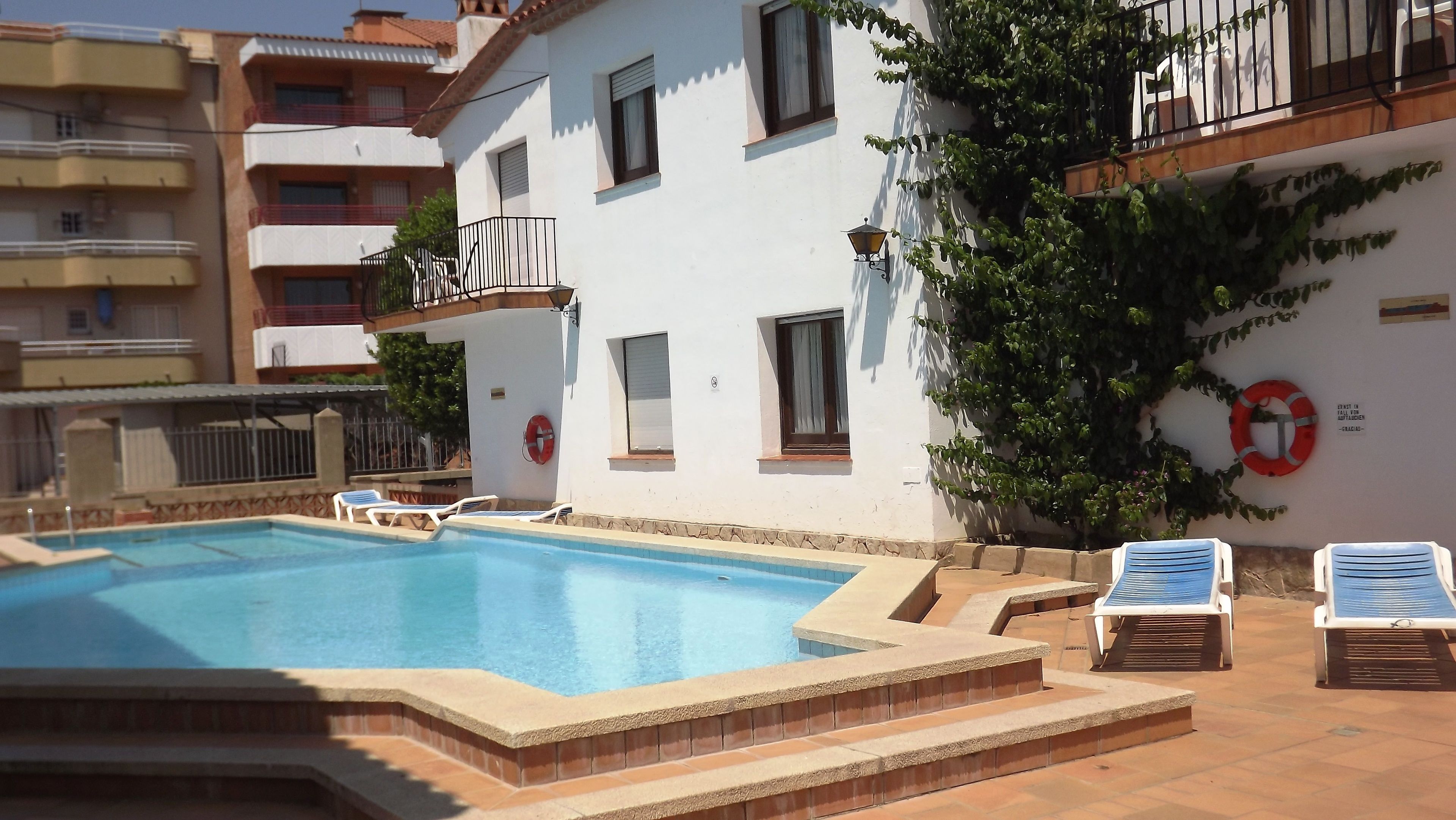 Una piscina con escalones de acceso, rodeada de sillas de descanso y un edificio blanco con balcones.