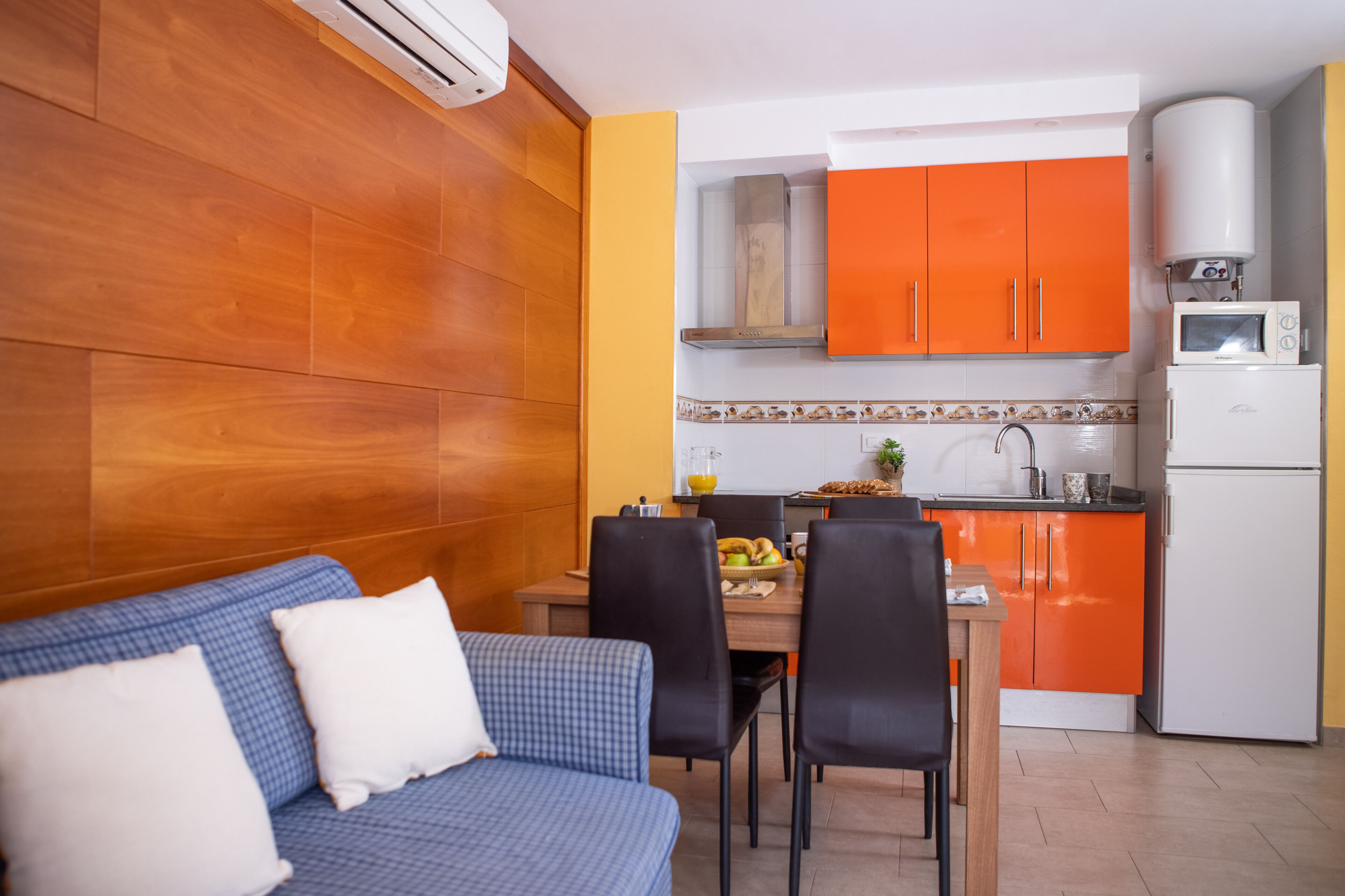 A compact living space with a kitchenette, a dining area with a blue sofa and black chairs, and a small kitchen with orange cabinets and white appliances.