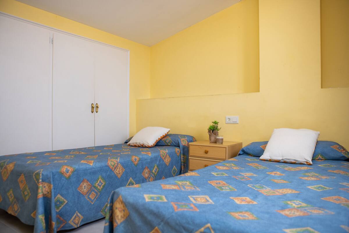 Ein Zimmer mit zwei Betten, jeweils mit einem gemusterten, blauen Bettüberwurf, einem Nachttisch aus Holz mit einer Pflanze und einem weißen Kleiderschrank.