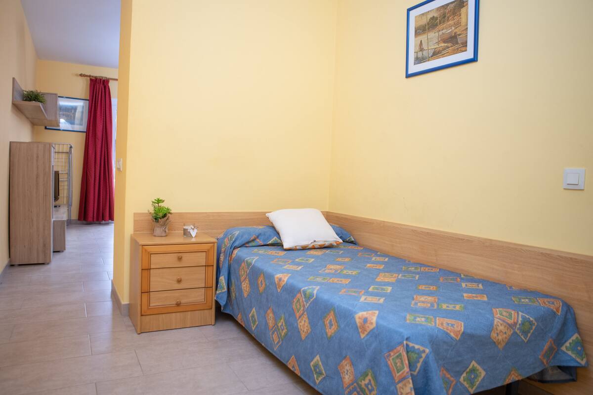 Ein Schlafzimmer mit einem Einzelbett, einem Nachttisch aus Holz, einer Kommode und einem roten Vorhang.