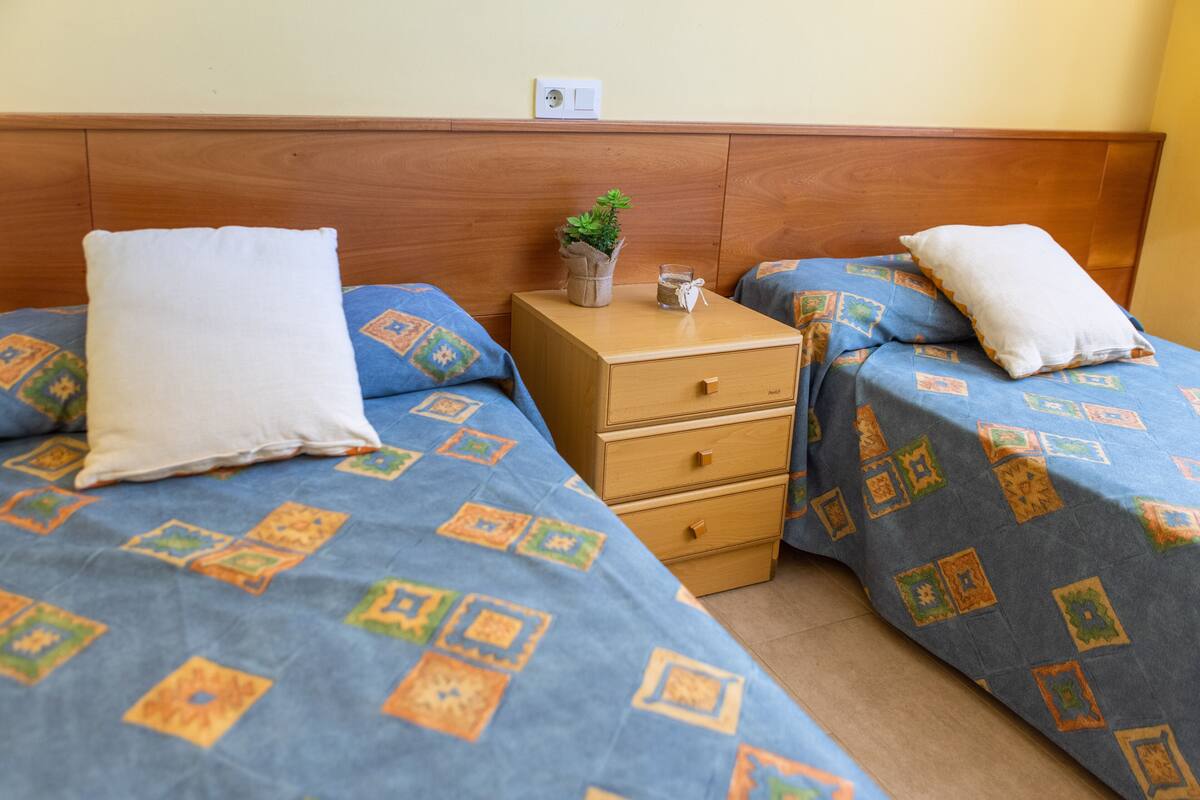Ein Schlafzimmer mit zwei Betten, jeweils mit einer blauen, gemusterten Bettdecke und weißen Kissen. Es gibt einen Holznachttisch mit einer kleinen Pflanze und einem Glas Wasser.