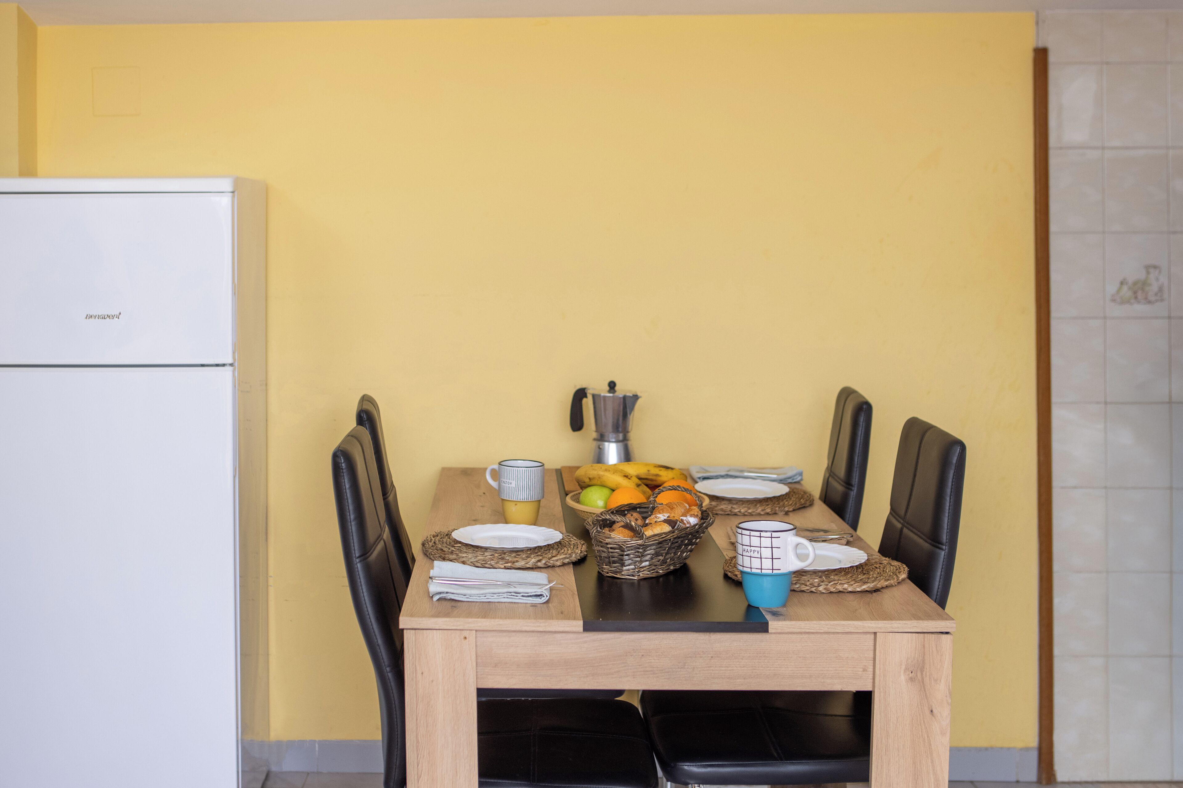Una mesa de comedor con sillas, una cesta de frutas y una cafetera.