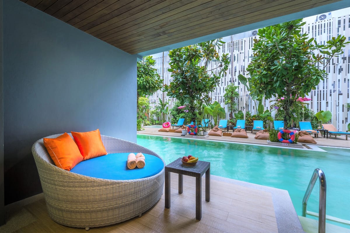 A poolside area with a wicker lounge chair, a small table with a bowl of fruit, and a view of the pool and surrounding greenery.