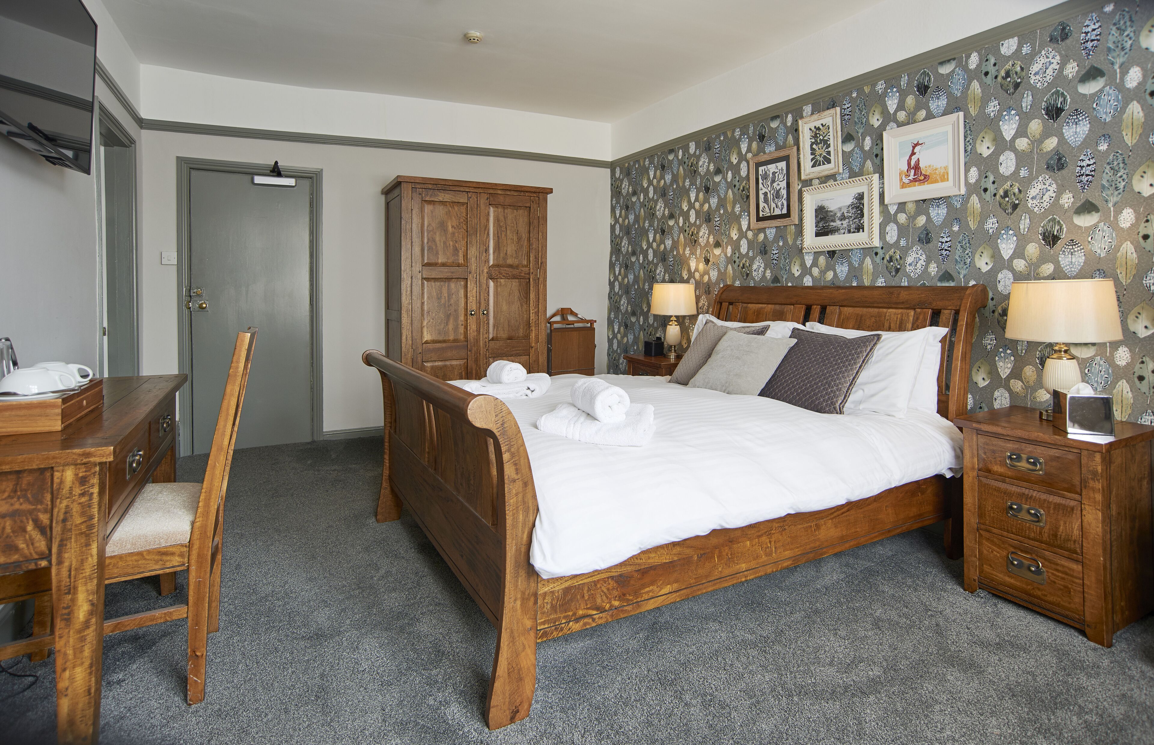 Une chambre à coucher avec un lit en bois, deux tables de chevet avec des lampes, une armoire en bois, un bureau avec une chaise et un sol recouvert d’une moquette.