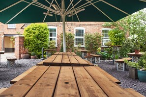 An outdoor patio area with wooden tables and benches, surrounded by potted plants and a brick building in the background. - Angel Inn Hotel (Pershore)