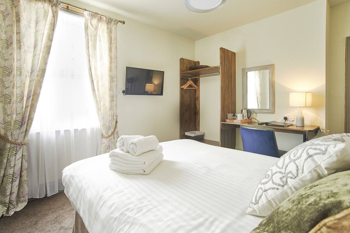 A hotel room with a bed, a desk with a chair, a mirror, a hanging coat rack, and a window with curtains.