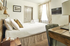 A hotel room with a bed, a desk, a chair, a bedside table, a lamp, a picture on the wall, and a window with curtains. - Angel Inn Hotel (Pershore)