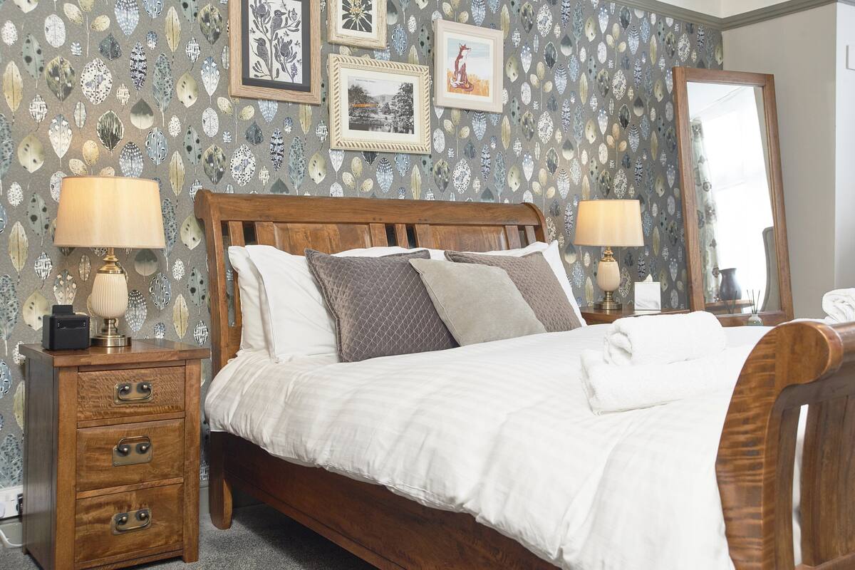 A bedroom with a wooden bed, white bedding, a nightstand with a lamp, and a patterned wallpaper.