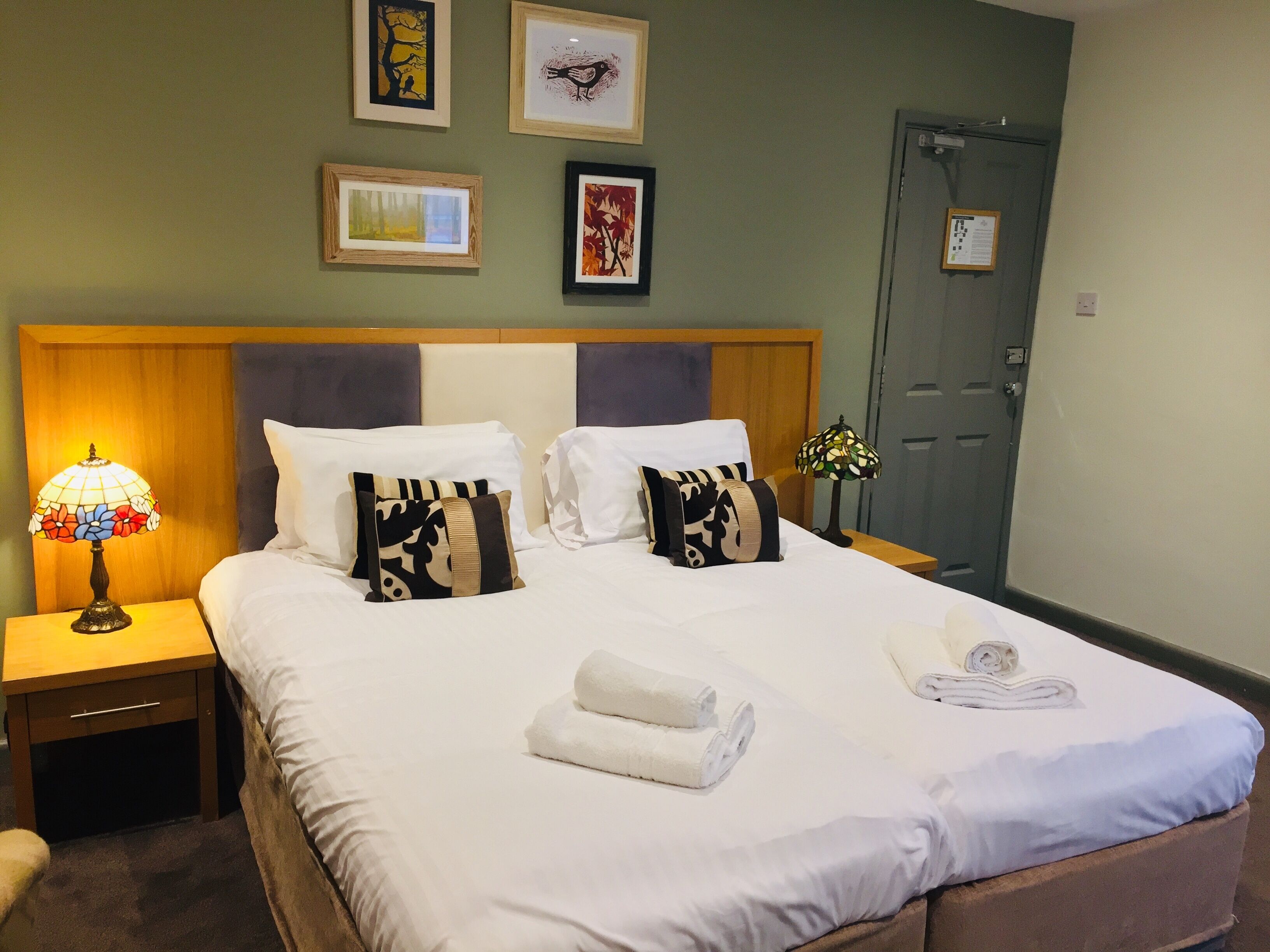 A neatly made bed with white linens, two pillows, and two rolled towels. There is a nightstand with a lamp and a framed picture on the wall above the bed.