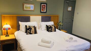 A neatly made bed with white linens, two pillows, and two rolled towels. There is a nightstand with a lamp and a framed picture on the wall above the bed.