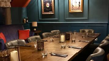 A dining area with a long wooden table set for six, surrounded by leather chairs. There is a chandelier above the table, and two framed awards on the wall.