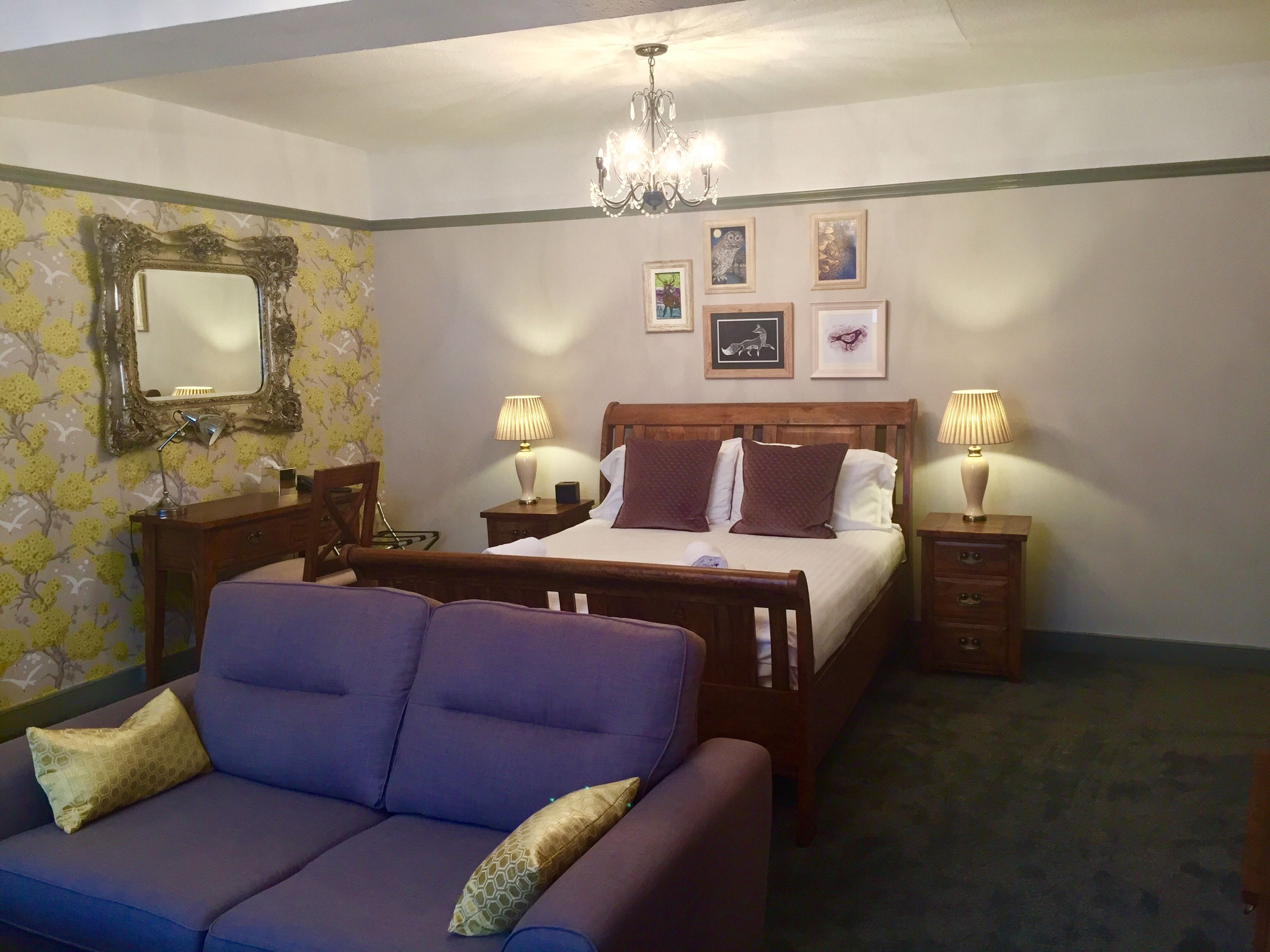 A bedroom with a bed, two bedside tables, a dresser, a purple sofa, a chandelier, and wall art.