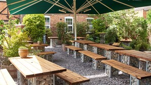 An outdoor patio with wooden tables and benches, surrounded by potted plants and a brick building in the background. - Angel Inn Hotel (Pershore)