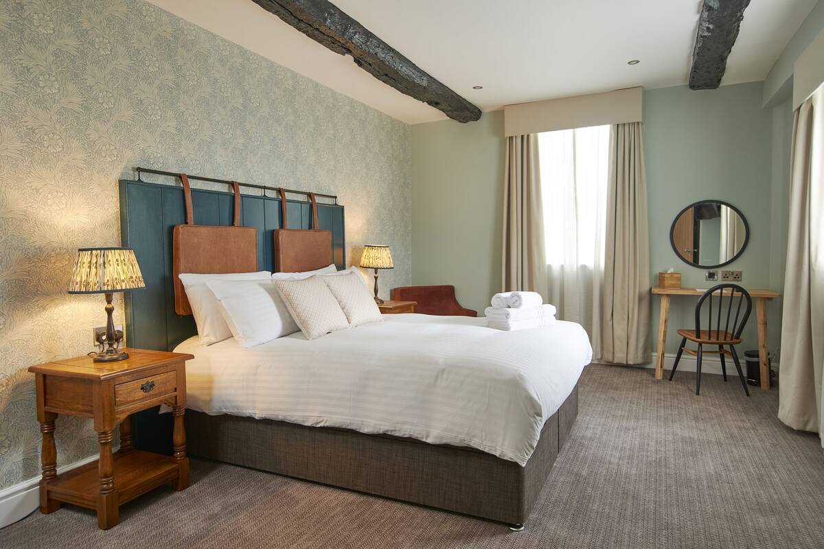 A hotel room with a large bed, two bedside tables with lamps, a wooden desk with a chair, a round mirror, and a window with curtains.
