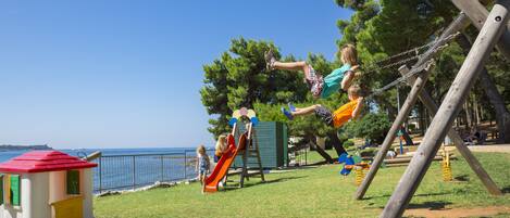 Un parque infantil con tobogán, columpios y una casita cerca del mar.