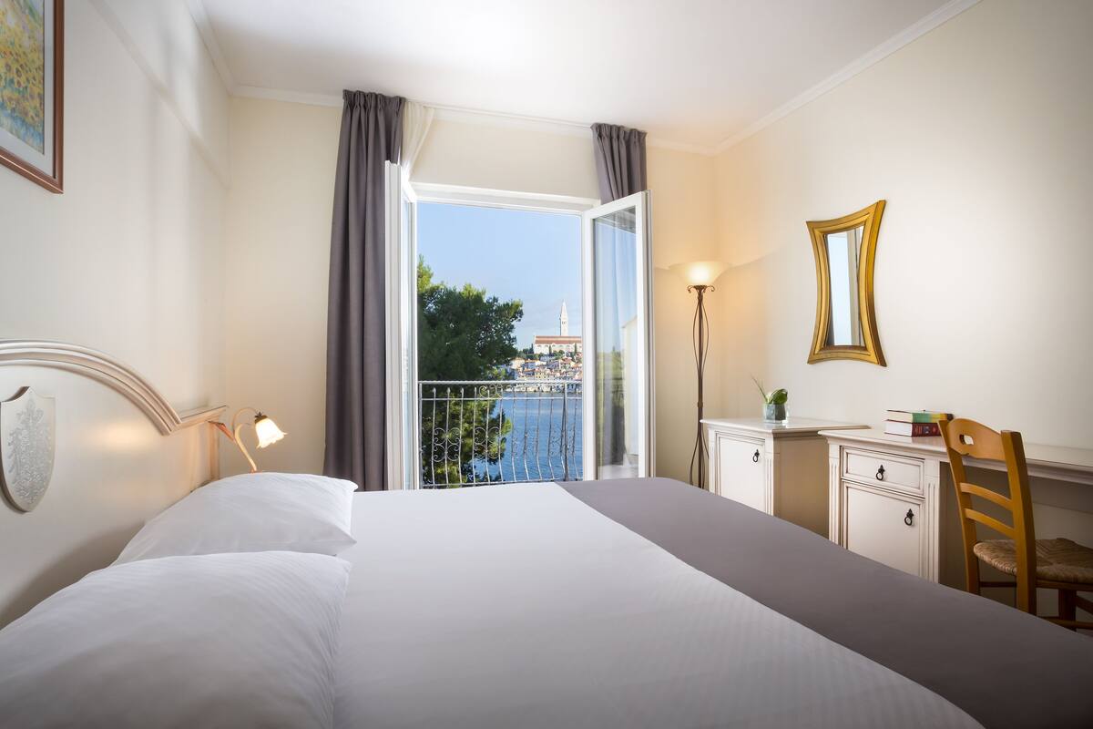 Habitación de hotel con una cama grande, un escritorio con silla, una lámpara y vista a un edificio desde una ventana abierta.