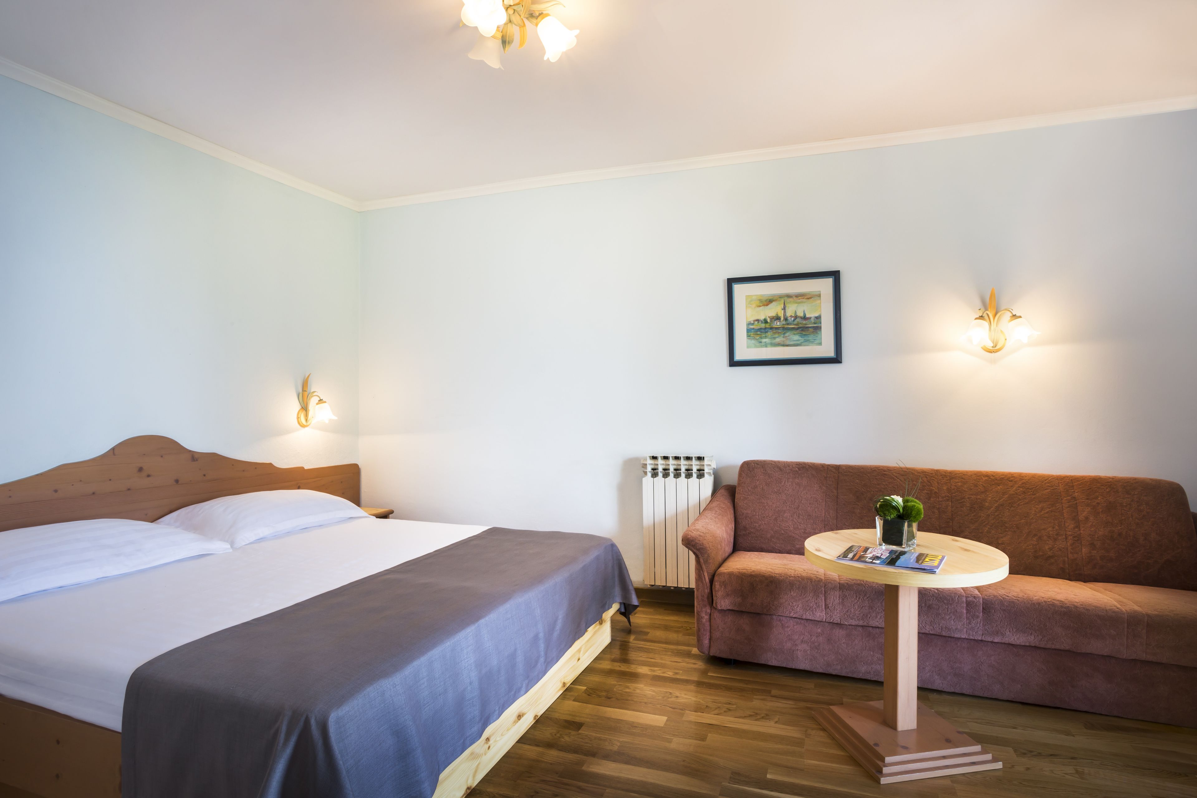 Habitación de hotel con cama, sofá, una mesita con una planta e iluminación en la pared.