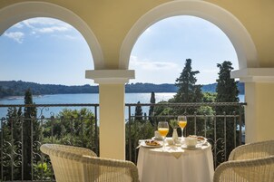 Un balcón con mesa lista para el desayuno, con vistas a un lago, rodeado de sillas de mimbre y barandilla de hierro forjado.