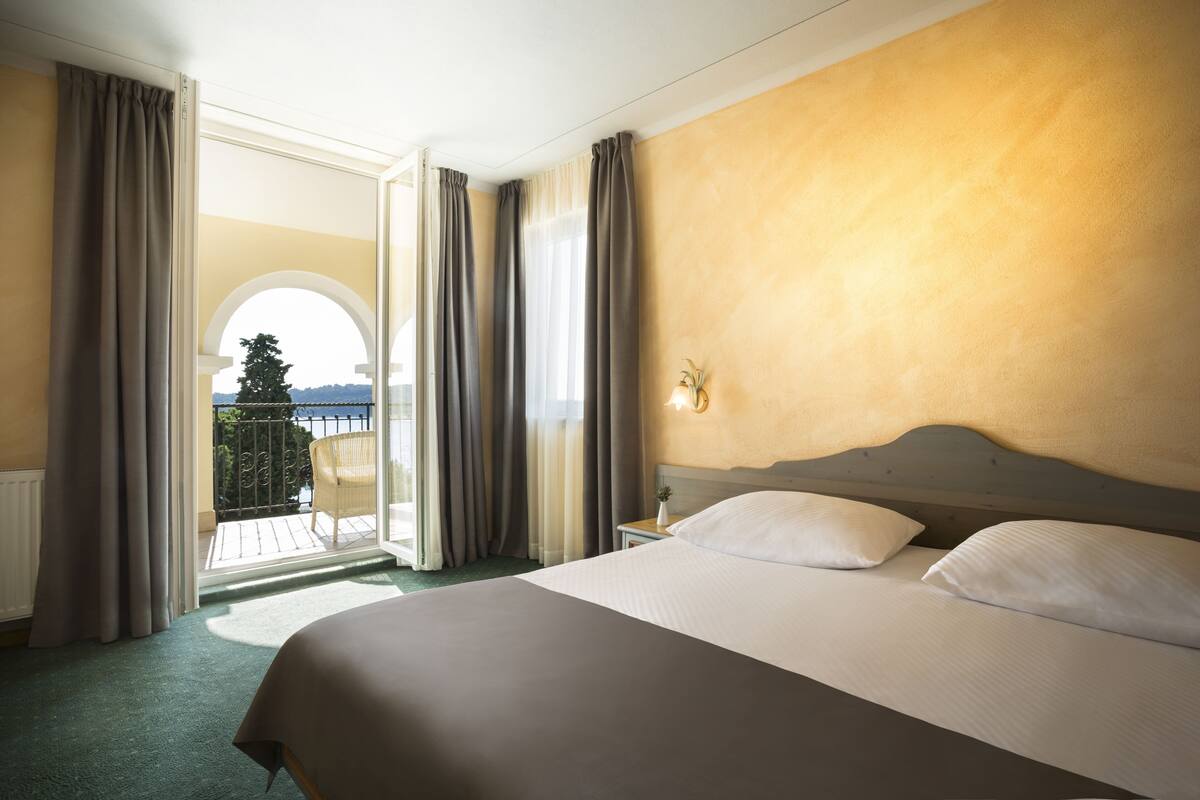 Habitación de hotel con una cama grande, una mesita de noche, una lámpara y vistas a un balcón con una silla y un árbol.
