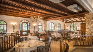 Restaurante com vigas de madeira, paredes de pedra e janelas em arco. Mesas montadas com toalhas brancas e velas.