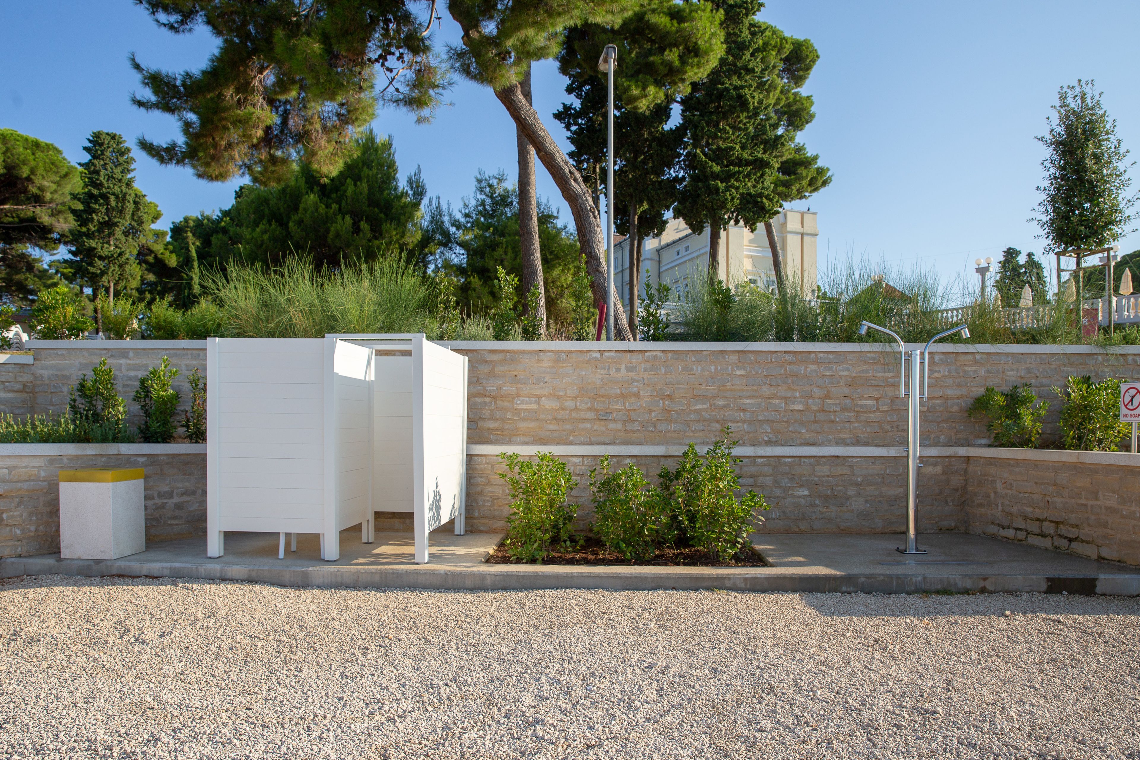 Un espacio exterior moderno con una mesa y sillas blancas, un contenedor de basura y una pequeña jardinera con plantas.