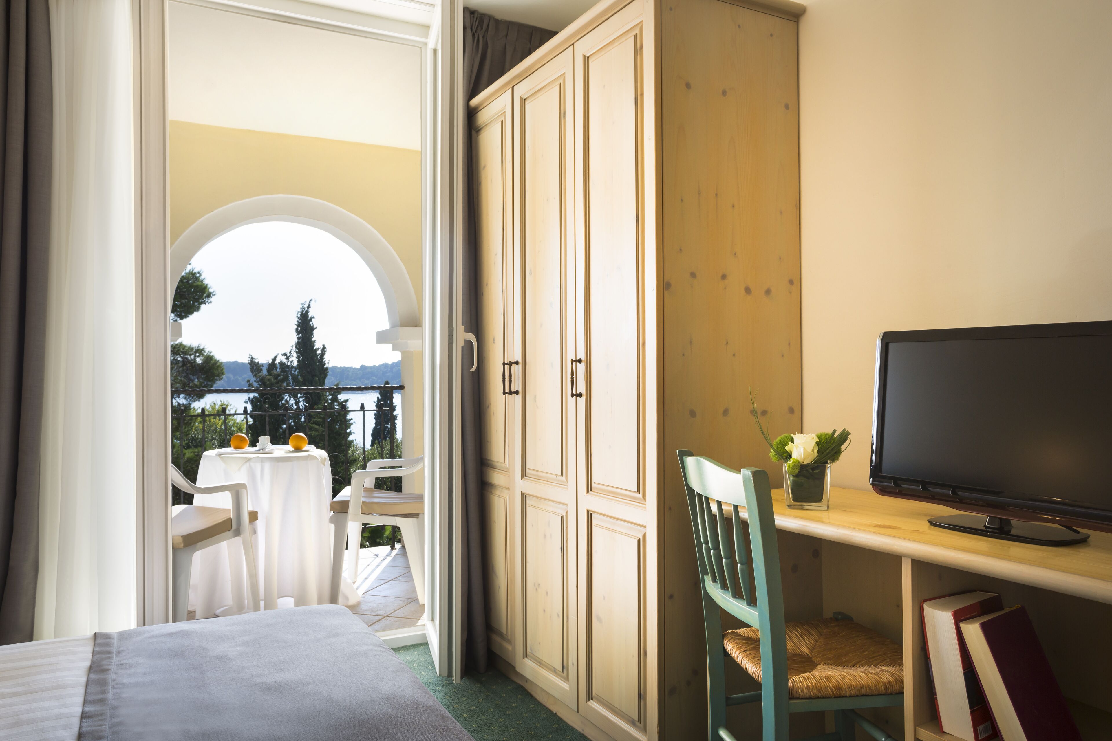 A room with a bed, a TV on a wooden shelf, a chair, and a table with a view of the outdoors.