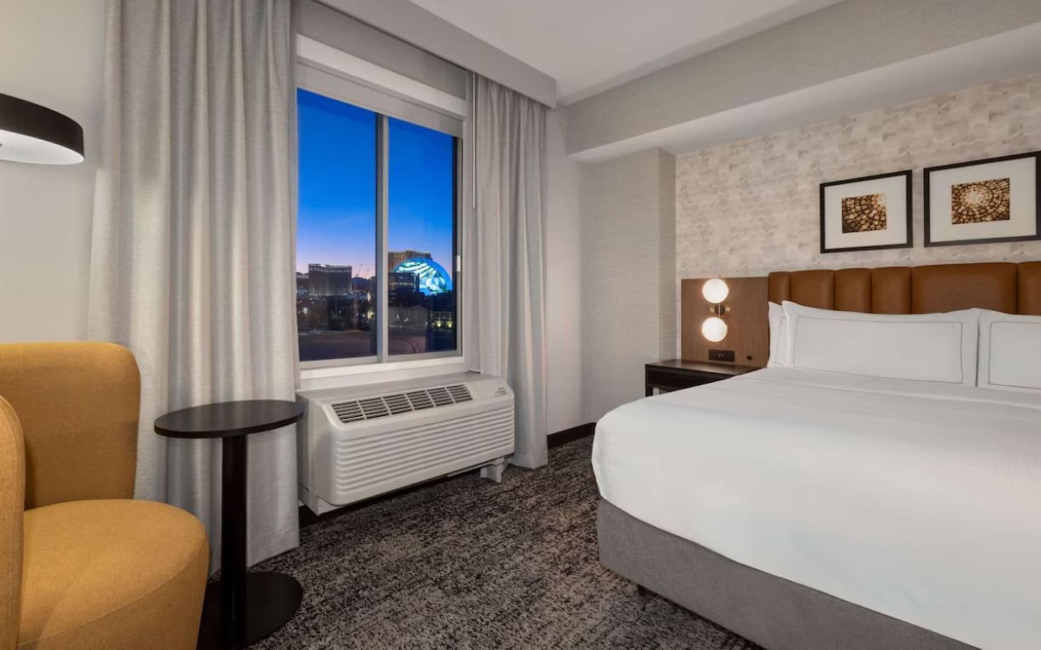 Una habitación de hotel con una cama grande, una silla, una mesita de noche con lámpara y una ventana con cortinas que ofrece vistas a la ciudad.