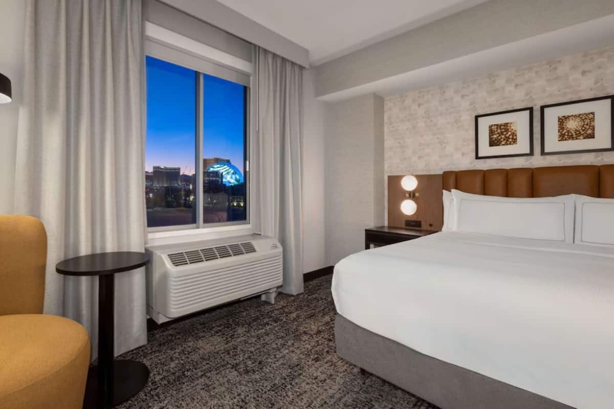 A hotel room with a large bed, a chair, a nightstand with a lamp, a window with curtains, and a city view.