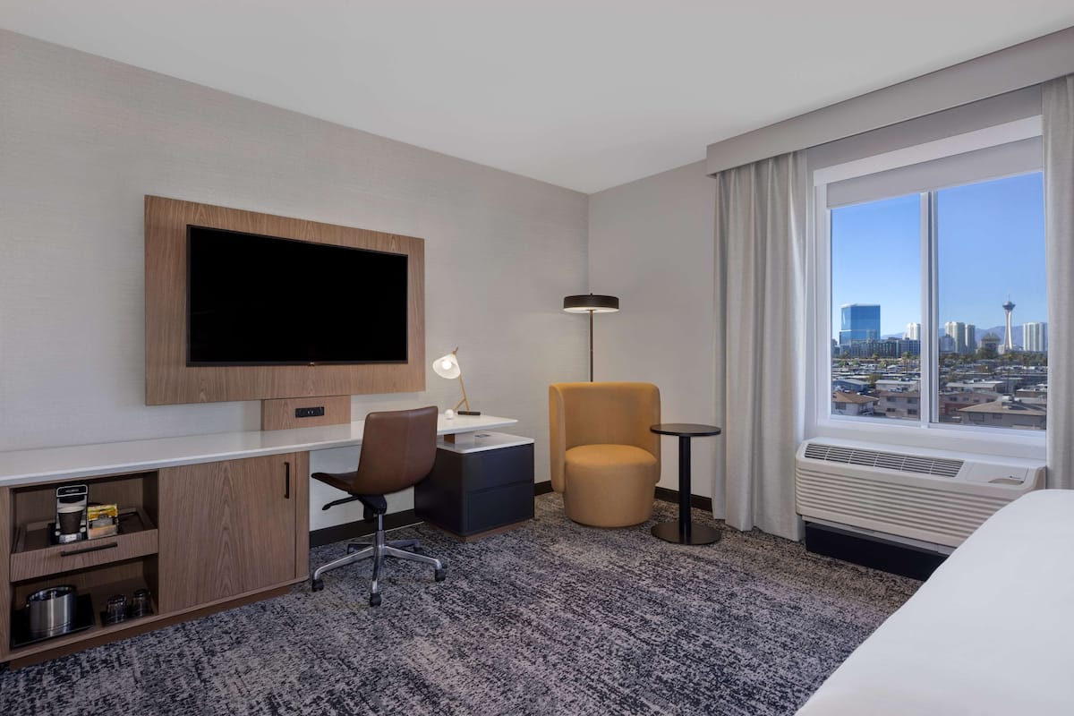 A hotel room with a large flat-screen TV, a desk with a chair, a small round table, a sofa, and a view of a cityscape through a large window.