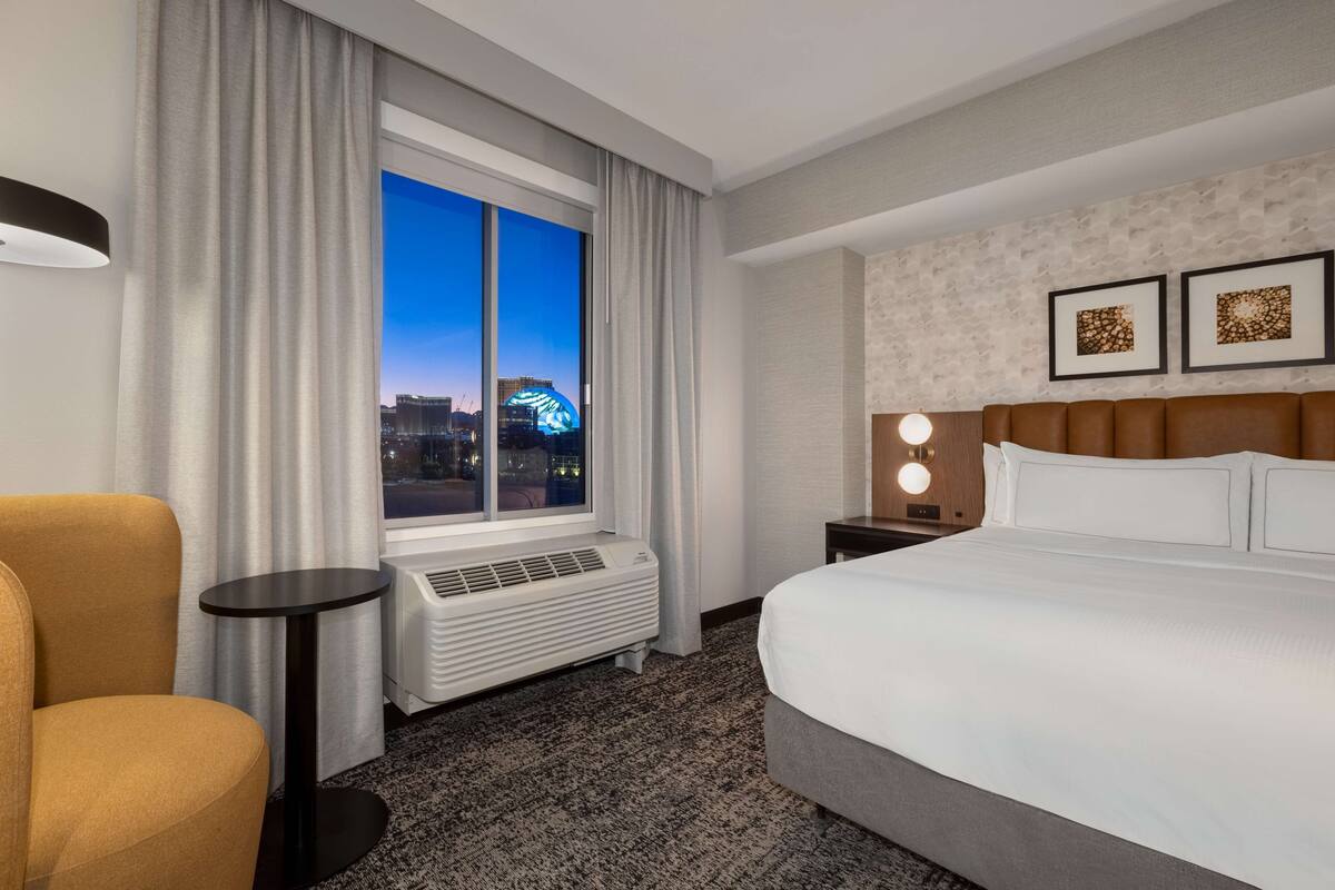 A hotel room with a large bed, a chair, a small table, a heater, and a window with a city view.