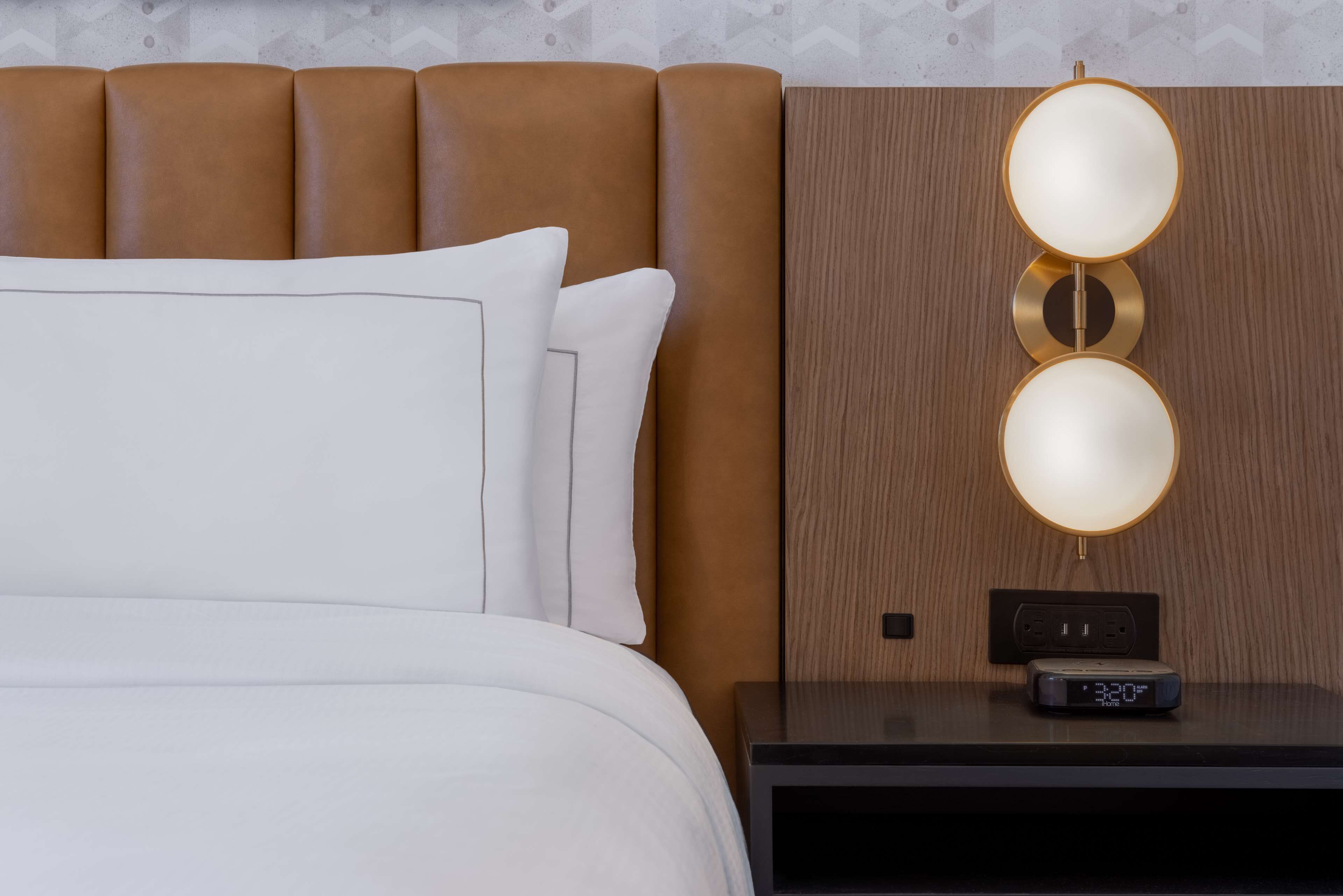 A neatly made bed with white linens and pillows, a wooden headboard, and a bedside table with a digital clock and a power outlet.