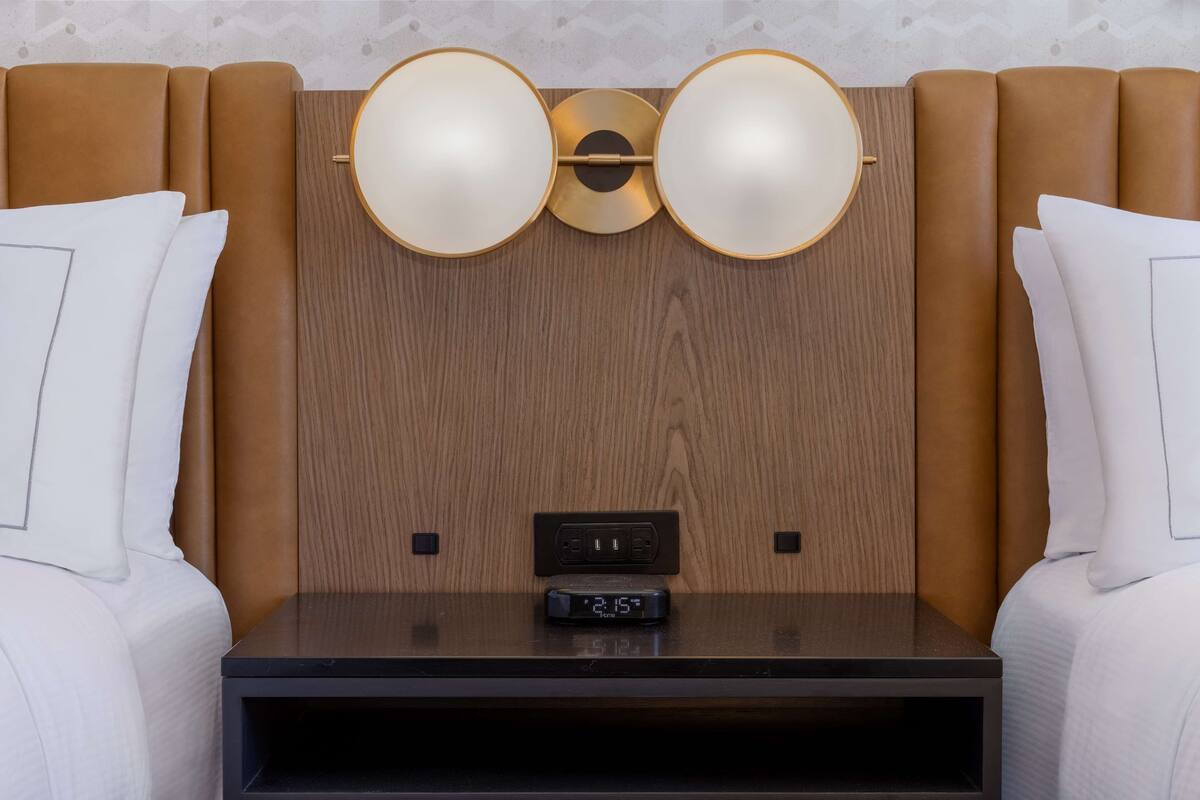 A hotel room with a wooden headboard, two bedside tables, a digital clock, and a wall-mounted light fixture.