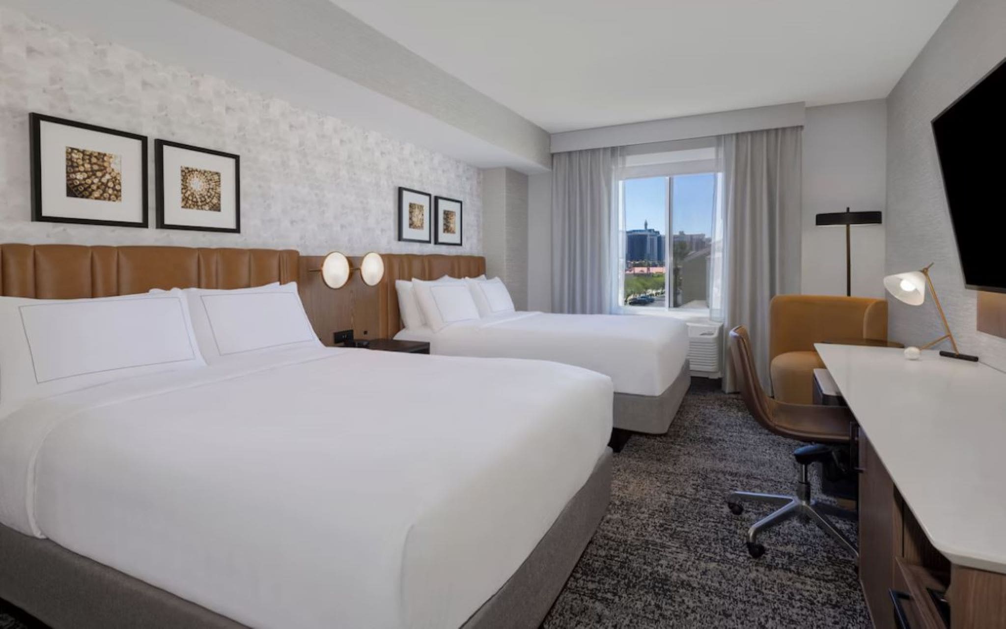 Habitación de hotel con dos camas, un escritorio, una silla, una lámpara y cuadros en la pared.