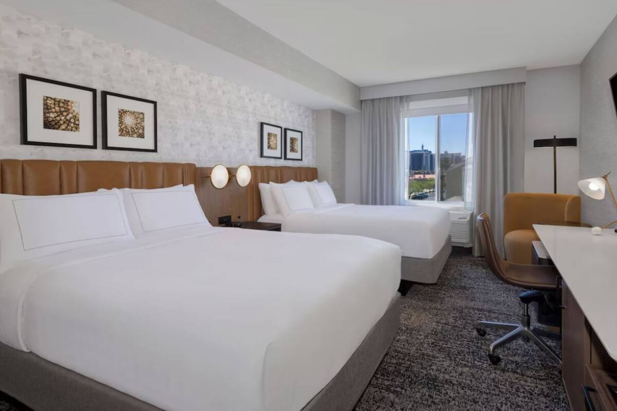 A hotel room with two beds, a desk, a chair, a TV, and a window with a city view.