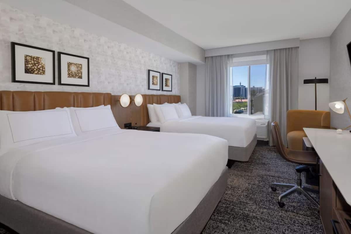 Une chambre d’hôtel avec deux lits, un bureau, une chaise, une lampe et des œuvres d’art encadrées au mur.