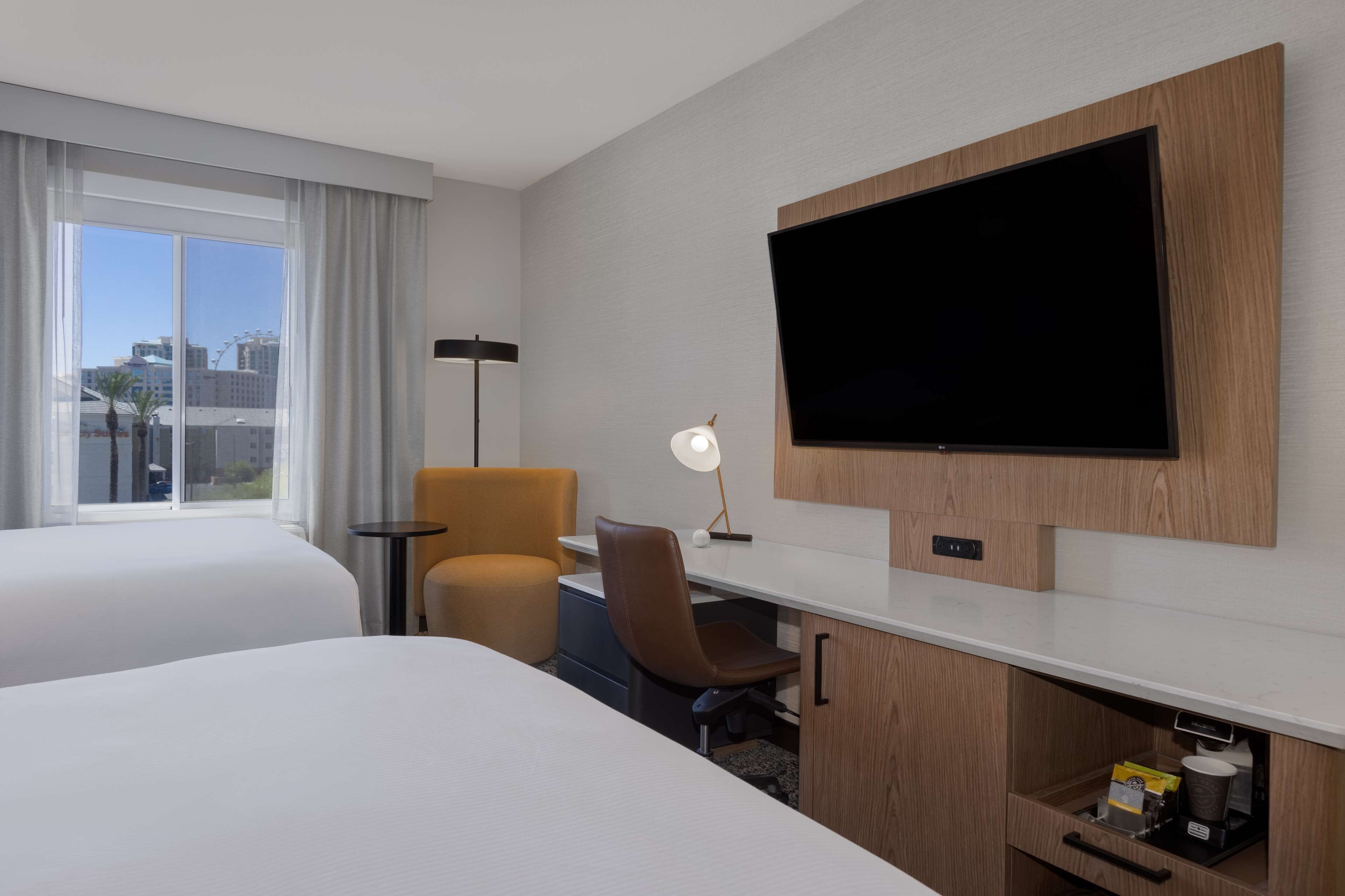 A hotel room with two beds, a desk with a chair, a television mounted on the wall, and a window with curtains.