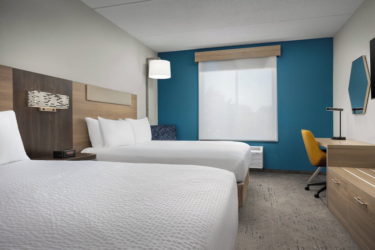 A hotel room with two beds, a desk, a chair, a lamp, a heater, and a large window.