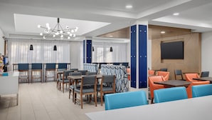 A modern dining area with tables and chairs, a chandelier, and a large screen on the wall. - Holiday Inn Express & Suites Greensboro Airport Area by IHG (Greensboro)